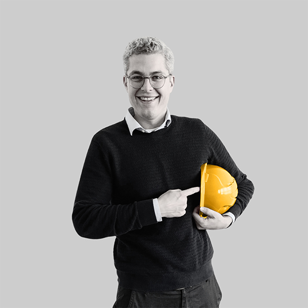 Team-rheform - Antoine Labrot, M.Sc. Wirtschaftsingenieur aus der nachhaltigen Immobilienentwicklung Gebäudeanalyse hält einen Bauhelm unter dem Arm, auf den er mit der anderen Hand zeigt